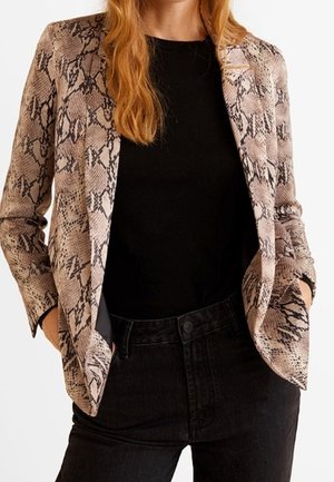 Vrouw draagt een beige en zwarte blazer met slangenhuidpatroon over een zwart shirt en zwarte hoge taille jeans, handen in de zakken.