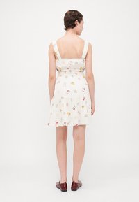 Vestido con estampado floral en una ligera tela color crema, con cintura fruncida, dobladillo con volantes y tirantes anchos. Combinado con zapatos burdeos brillantes.