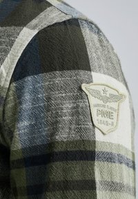 Chemise en flanelle avec un motif à carreaux dans des tons de vert, bleu marine et gris, avec un tissu texturé et un patch logo de couleur crème sur l'épaule.