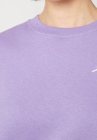 Sudadera lavanda hecha de una tela suave, con un cuello acanalado y un pequeño logo blanco en el pecho. Textura suave, ajuste casual.