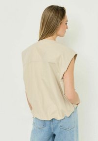 Donna con lunghi capelli biondi che indossa un top beige senza maniche e jeans azzurri, in piedi davanti a uno sfondo bianco semplice.