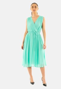 Un vestido midi sin mangas, plisado, en un tono teal claro con un diseño cruzado, cintura ajustada y un lazo ajustable. Unas sandalias de tacón negro completan el look.