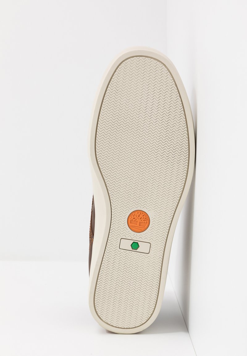 Braune Schuhsohle mit wellenförmigem Muster, ausgestattet mit einem orangefarbenen und grünen Logo sowie Akzenten. Der Rand ist cremefarben und hat ein geomtrisches Profil.