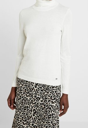 Femme portant un pull à col roulé blanc uni et une jupe imprimée léopard noir et blanc, debout devant un fond gris uni.