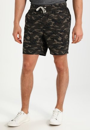 Homme portant un short camouflage noir et gris avec un cordon blanc et des baskets blanches, debout les jambes légèrement écartées.