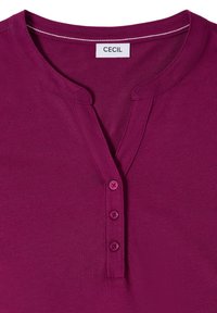Chemise couleur prune avec un col rond, trois boutons assortis sur la patte de boutonnage avant, et une étiquette blanche "CECIL" à l'intérieur du col.