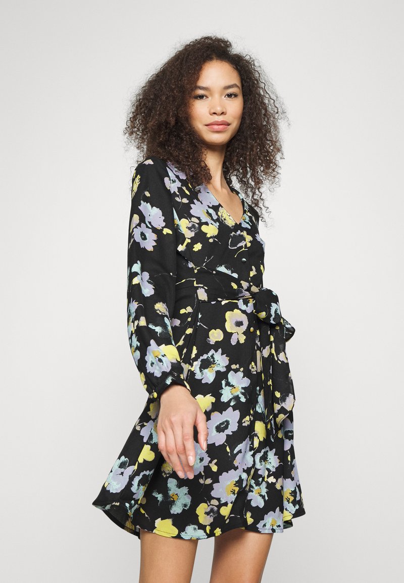 Vestido preto floral com mangas compridas, apresentando um design envolvente, decote em V e uma cintura atada. Flores em amarelo, lavanda e verde-água criam um padrão vívido.