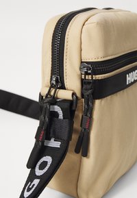 Borsa in nylon beige con due scomparti con zip, tracolla nera con testo bianco e dettagli di marca sulle zip.