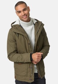 Olijfgroene jas met een bontgevoerde capuchon, knoopsluiting en zijzakken; gedragen over een lichtgrijze, getextureerde coltrui.