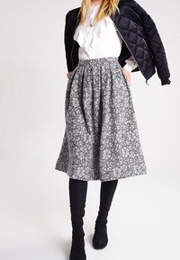 Jupe midi plissée grise avec motif floral, en tissu léger, associée à un chemisier blanc et une veste bomber matelassée noire.