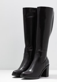 Paire de bottes montantes en cuir noir avec talons carrés et fermetures éclair latérales, présentées sur une surface blanche avec un fond neutre.