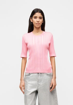 Donna con capelli lunghi e scuri indossa un maglione rosa a maniche corte a coste e pantaloni larghi in tessuto metallico grigio-argento su uno sfondo semplice.