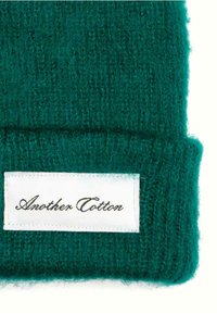 Zelená pletená čiapka s textúrovaným, jemným povrchom a prehnutým lemem. Obsahuje bielu nálepku s nápisom "Another Cotton" písaným kurzívou.