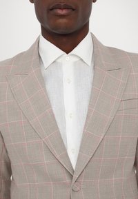 Šviesiai pilkas lino švarkas su rausvu plaid raštu, derinamas su baltu lino marškiniu, kuriame yra smailas apykaklė ir mygtukų uždarymas.