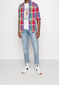 Czerwona, niebieska i zielona koszula w kratę noszona na białej koszulce z grafiką, połączona z jasnoniebieskimi dżinsami i białymi butami sportowymi z granatowymi akcentami.