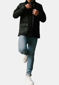 Giacca nera con bottoni, dotata di due tasche frontali, indossata su una camicia scura, abbinata a jeans azzurri chiari e scarpe da ginnastica bianche.