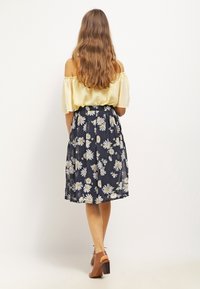 Top giallo off-the-shoulder abbinato a una gonna plissettata blu scuro con margherite bianche. Sandali con tacco marroni completano l'outfit.