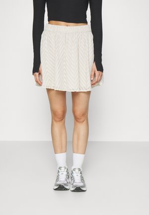 Licht beige mini-rok met een visgraatpatroon, een elastische tailleband en een gestructureerde afwerking, gecombineerd met witte sokken en zilveren sneakers.