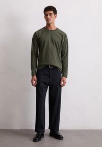 Olivgrünes Langarmshirt mit lockerer Passform, kombiniert mit dunkelblauen, gefalteten Hosen und schwarzen Schuhen. Einfaches Design, glatte Textur.