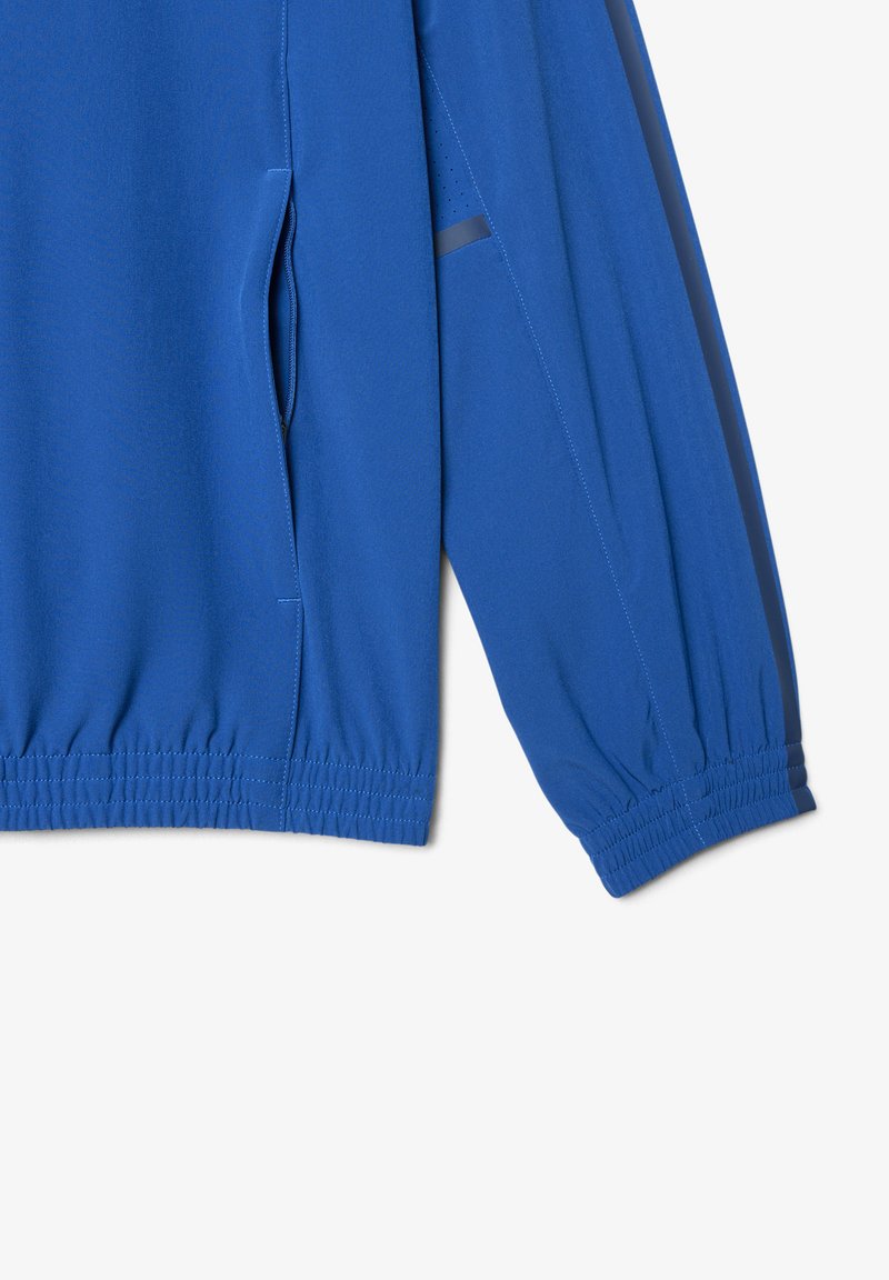 Blauer Jackenärmel und Seitenansicht mit elastischem Bund und sichtbarem Taschenreißverschluss auf weißem Hintergrund.