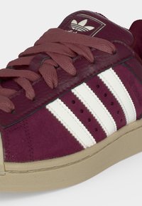 Μπορντό sneaker Adidas με λευκές τρεις ρίγες, υφασμένο δέρμα, κορδόνια και μπεζ σόλα, που δείχνει το λογότυπο της Adidas στη γλώσσα.