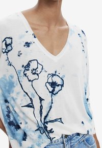 Pull en tricot blanc avec un motif floral bleu et un col en V. Le tissu a une texture douce avec une coupe décontractée.