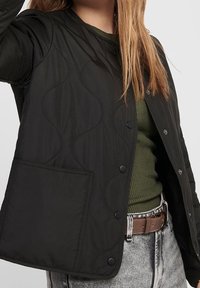 Sort quiltet jakke med rund halsudskæring, trykknaplukninger og en enkelt lomme. Båret over en grøn top og grå højtaljede jeans.