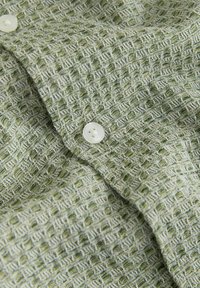 Gros plan sur un tissu vert clair texturé avec des fils blancs et deux boutons blancs sur un vêtement plié.