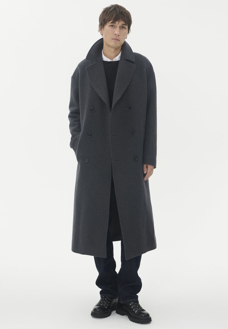 Ung man som står i en lång mörkgrå dubbelknäppt kappa, svarta byxor, svarta stövlar och vit skjorta med svart tröja, neutralt bakgrund.