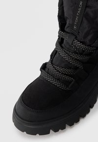 Black ankle boot featuring a combination of smooth leather and textured suede, with a thick, patterned lace and a rugged, serrated sole.
