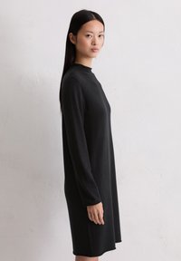 Schwarzes langärmliges Kleid mit hohem Kragen, aus weichem Stoff gefertigt. Verfügt über einen geraden Schnitt und reicht bis zur Mitte des Oberschenkels, minimalistisches Design.