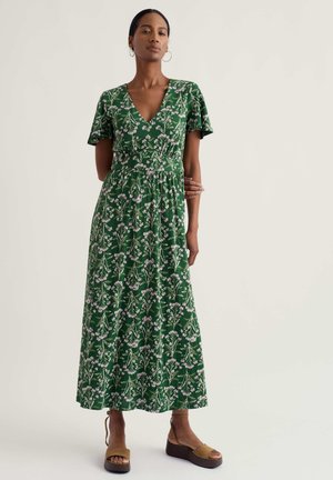 Vrouw in een groene midi-jurk met bloemenprint, V-hals en korte zwierige mouwen, met beige platform sandalen, staand tegen een effen achtergrond.