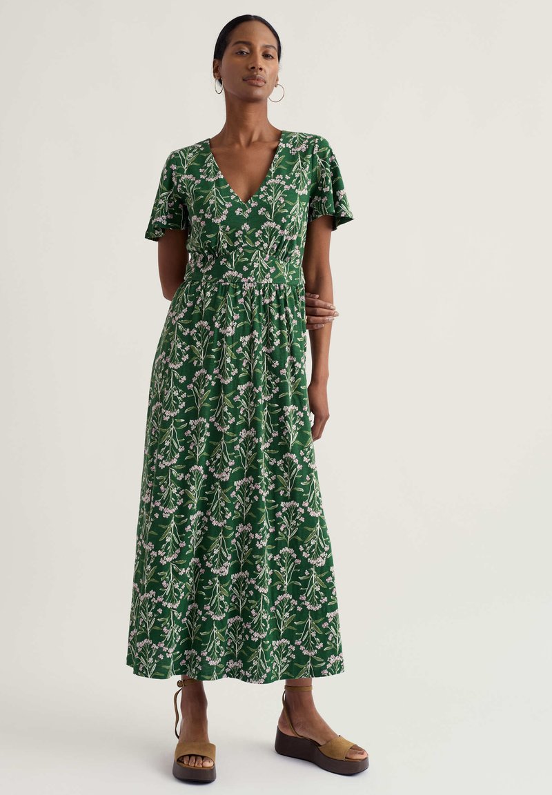 Femme portant une robe mi-longue à fleurs vertes avec un col en V et des manches courtes flottantes, ainsi que des sandales compensées couleur cuir, se tenant devant un fond uni.