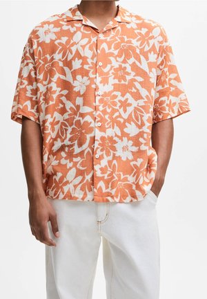 Hombre vistiendo una camisa de botones de manga corta con flores en naranja y blanco, combinada con pantalones blancos, de pie con las manos en los bolsillos.