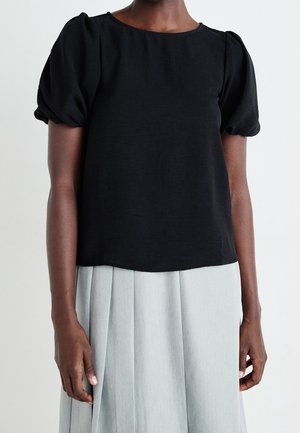 Musta, lyhythihainen pusero, jossa on pussihihat; sileä kangas ja pyöreä kaula-aukko. Yhdistetty vaaleanharmaaseen plisseerattuun hameeseen.