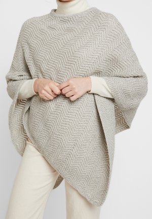 Osoba ubrana w jasnobeżowy, teksturowany, dziany ponczo na kremowym swetrze z golfem i kremowych spodniach, ręce delikatnie złożone na wysokości talii.