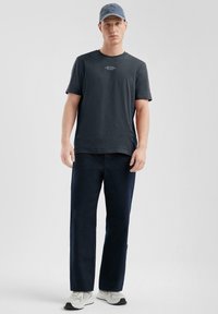 T-shirt à manches courtes gris foncé avec du texte blanc, associé à un pantalon bleu marine. Le modèle porte une casquette en denim bleu clair et des baskets grises.