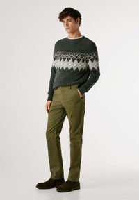 Tmavosivý sveter s geometrickými vzormi v bielej, oranžovej a čiernej farbe, spárovaný s olivovozelenými nohavicami a tmavos hnedými semišovými čižmami.