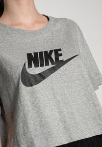 Šedé zkrácené tričko s černým logem Nike na přední straně. Vyrobeno z měkké látky s kulatým výstřihem a krátkými rukávy.