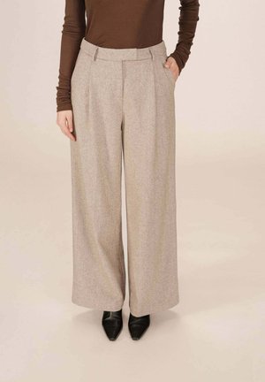 Femme portant un pantalon large beige, un haut à manches longues marron et des chaussures à bout pointu noires, se tenant avec une main dans la poche.