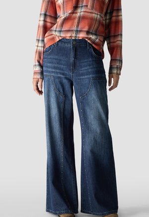 Person wearing wide-leg dark blue jeans with studded detailing and an orange and white plaid shirt tucked in at the waist.