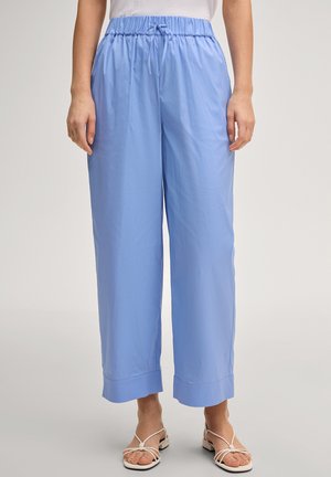 Frau trägt lockere hellblaue Hosen mit elastischem Bund und Kordelzug, kombiniert mit weißen flachen Sandalen mit Riemchen.