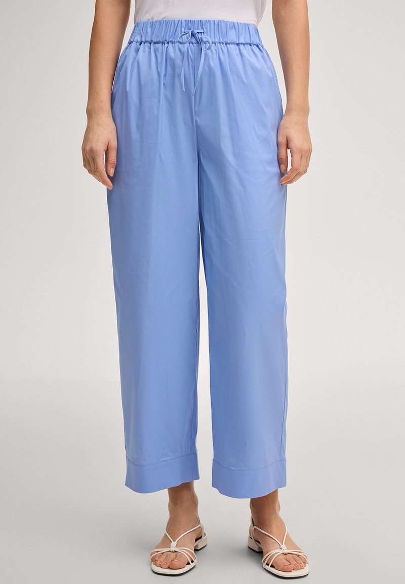 Femme portant un pantalon large bleu clair avec taille élastique et cordon de serrage, associé à des sandales blanches plates à brides.