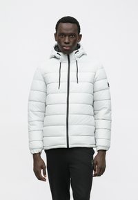Λευκό μπουφάν τύπου puffer με οριζόντια ραφή, μαύρο φερμουάρ και ρυθμιζόμενη κουκούλα με κορδόνι. Φορεμένο πάνω από σκούρα παντελόνια.