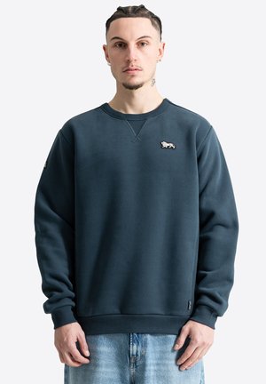 Mann mit geflochtenem Haar, der einen dunkelblauen Sweatshirt mit Rundhalsausschnitt und kleinem Löwenemblem auf der linken Brust sowie hellblaue Jeans trägt.