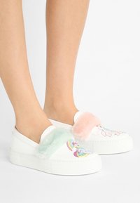 Białe buty wsuwane z kolorowymi wzorami jednorożców, ozdobione puszystymi pastelowymi akcentami w kolorze mięty i różu na paskach. Grube białe podeszwy.