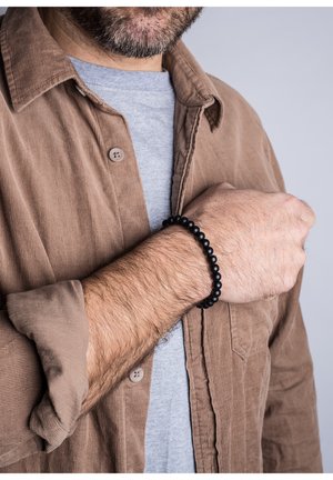 Pulsera de cuentas negras en la muñeca, hecha de cuentas redondas lisas. Llevada con una camisa de pana marrón y una camiseta gris debajo.