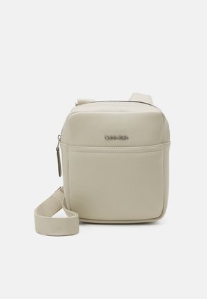 Kompakte beiges Umhängetäschchen mit Vordertasche, Reißverschluss, verstellbarem Stoffriemen und kleinem Metall-Logo von Calvin Klein auf der Vorderseite.