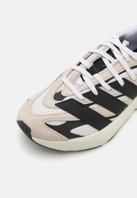 Beige and black sneaker with mesh and suede materials, featuring white laces, three black stripes, and a textured rubber sole.