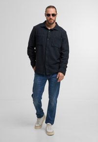 Dunkelblaues Hemd mit Knöpfen und zwei Brusttaschen, getragen über hellblauen Jeans und beigen Sneakers. Glatte Stoff mit strukturierter Oberfläche.